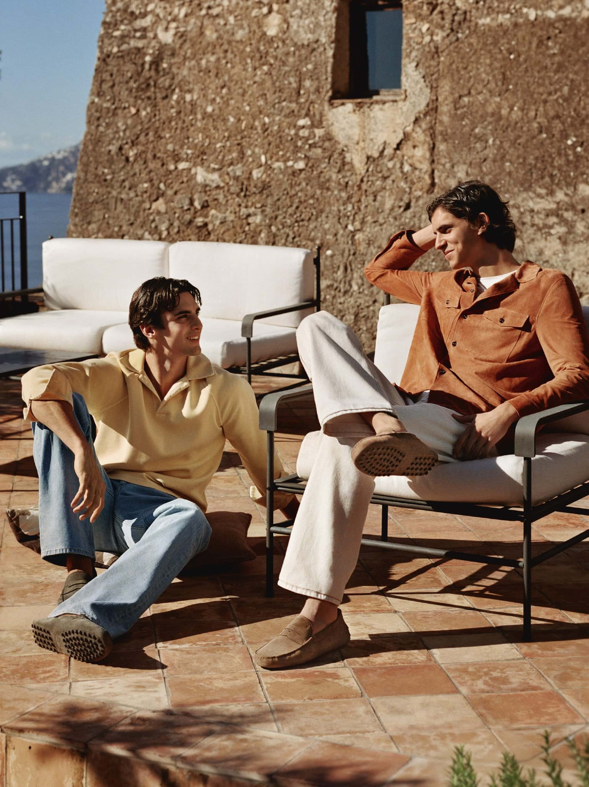Two adult men in casual attire relaxing on outdoor furniture on a terracotta tiled patio, with stone wall and seascape backdrop.