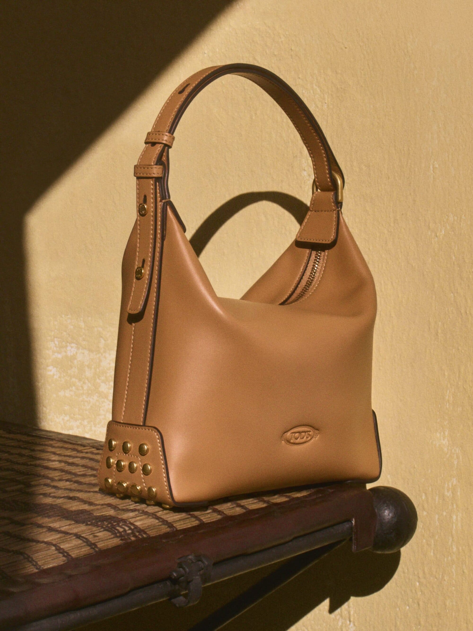 Tan leather Tod's hobo bag with studded detail and handle resting on a dark wood surface against a yellow wall with sunlight shadow.
