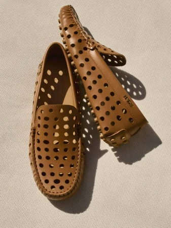 Two tan leather driving shoes with perforated details are angled on a textured light background, casting shadows and showing the 'Tod's' logo.
