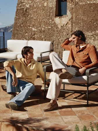 Two men relaxing outdoors on a tiled terrace with a rustic stone wall background for Tod's Spring 2026 ad campaign.