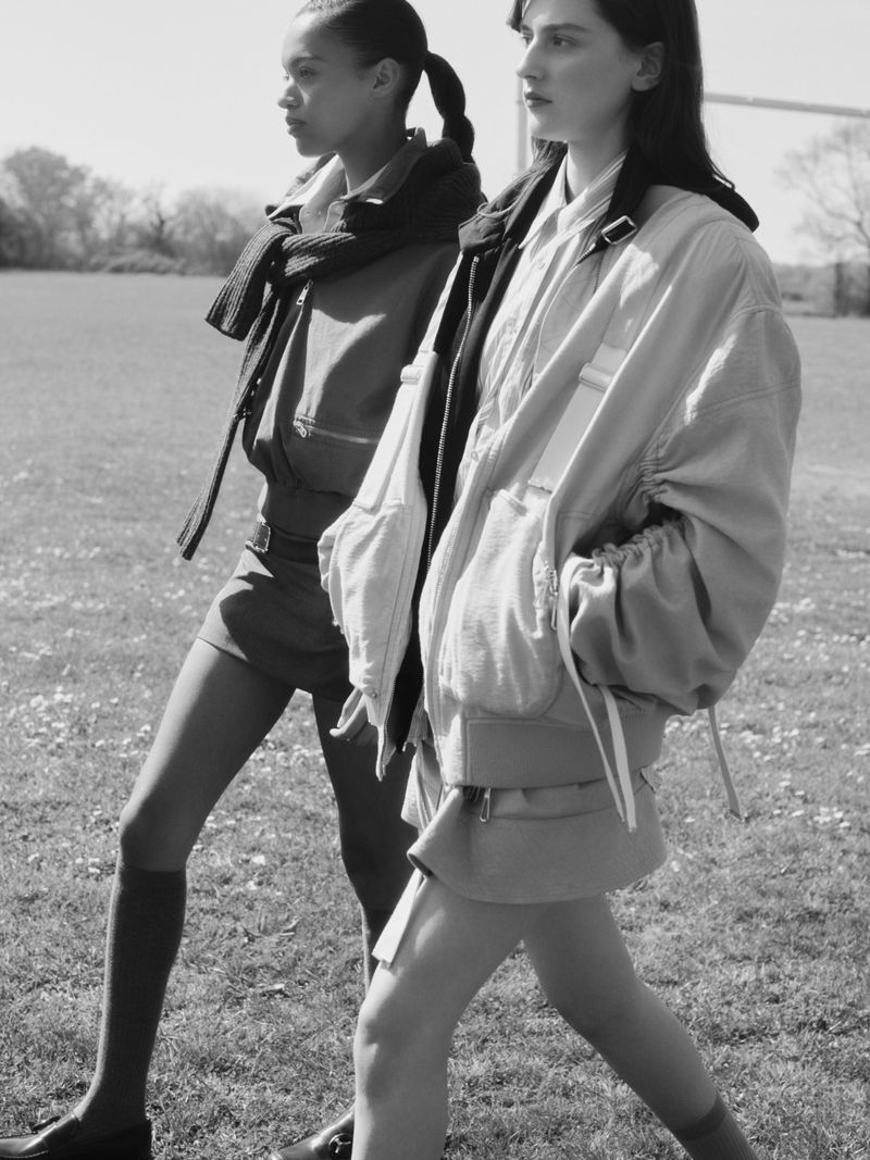 Two women walk side-by-side on a grassy field wearing bomber jackets and skirts, lit by natural light in a monochrome photograph.