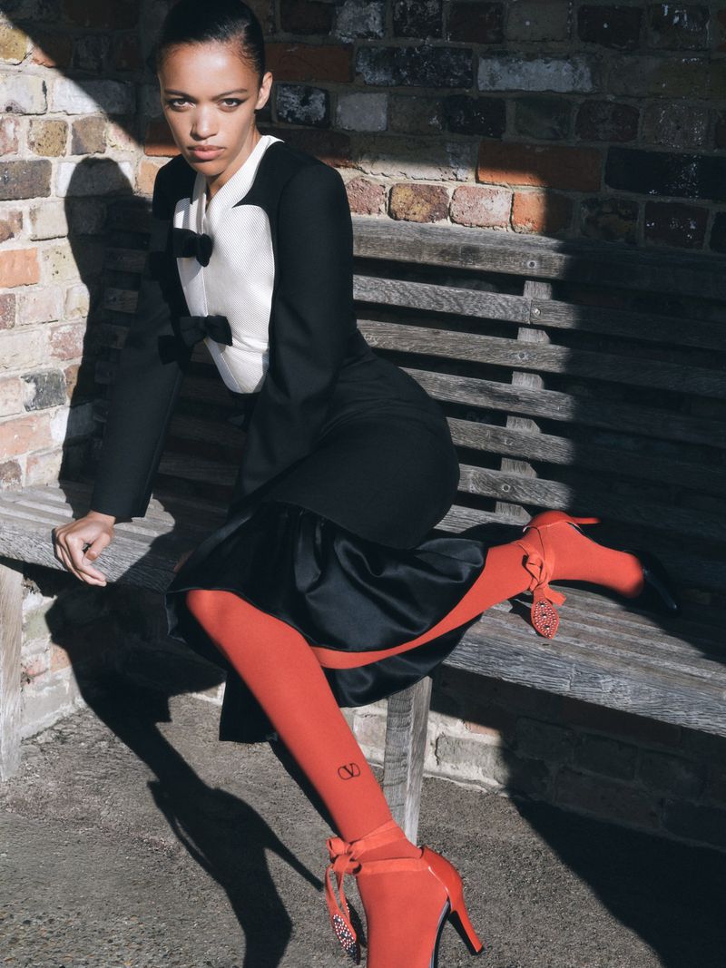 Woman in black and white outfit and red tights sitting on bench in outdoor setting, posing in natural light. Fashion editorial.