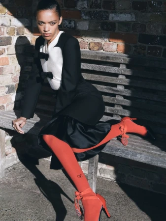 Woman in black and white outfit and red tights sitting on bench in outdoor setting, posing in natural light. Fashion editorial.
