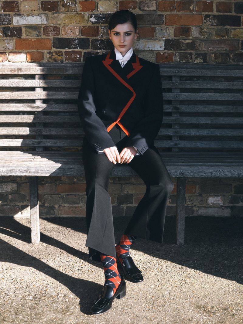 Young woman in a black suit with red trim poses on a wooden bench in an outdoor setting with a brick wall backdrop.