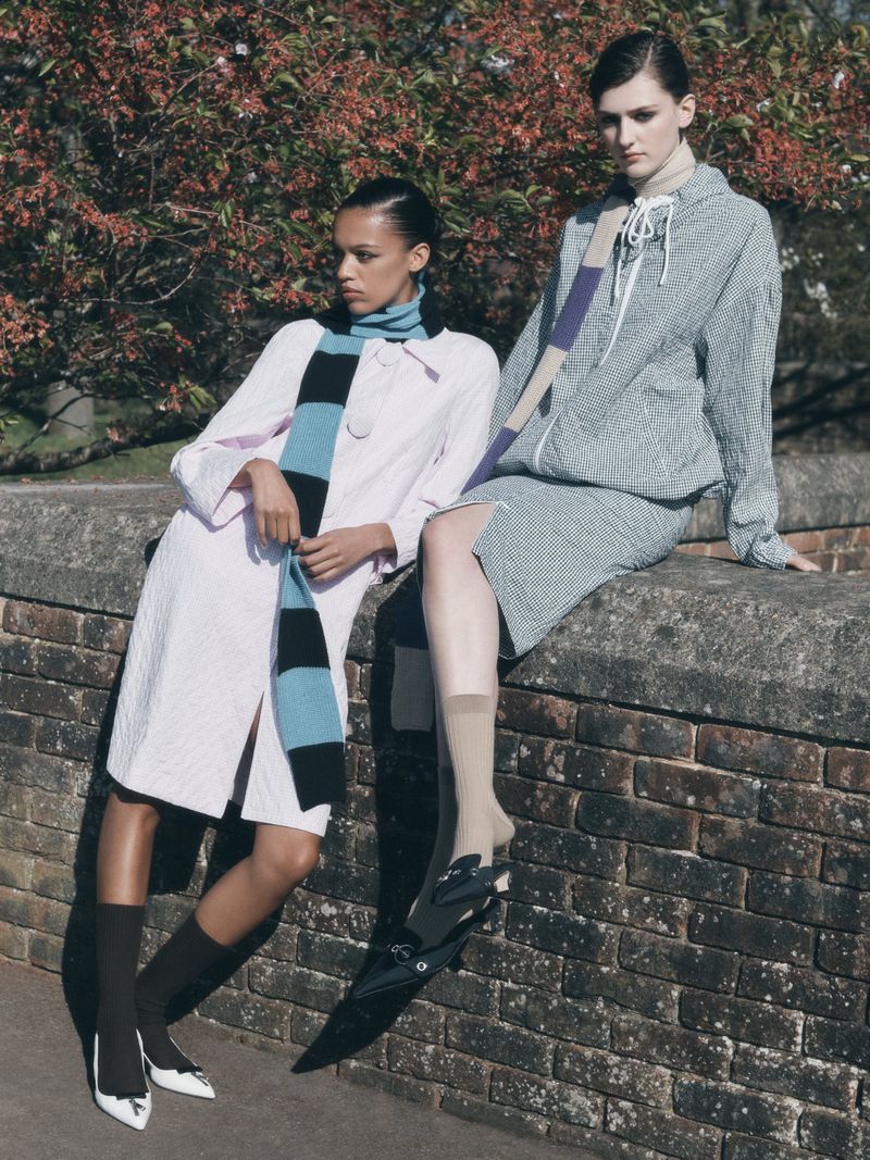 Two women in stylish coats and scarves pose outdoors against a brick wall with a backdrop of budding trees in the background.