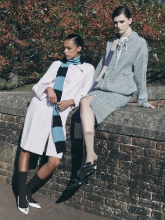 Two women in stylish coats and scarves pose outdoors against a brick wall with a backdrop of budding trees in the background.