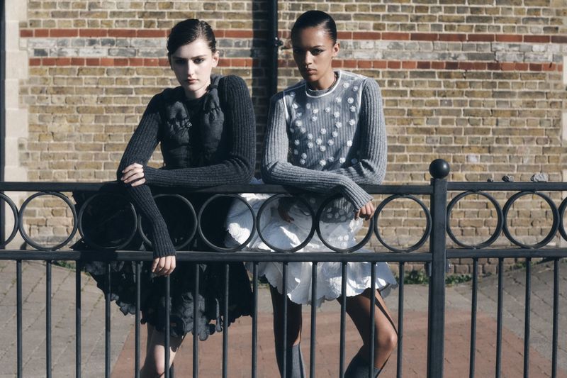 Two fashion models in gray sweaters and white skirts pose in front of a brick wall and black metal fence in an outdoor setting.