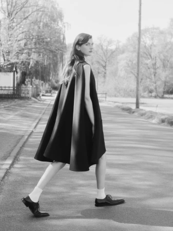 Monochrome fashion shot of a young woman in a black dress walking on a road, with trees in the background, wearing socks and leather shoes.