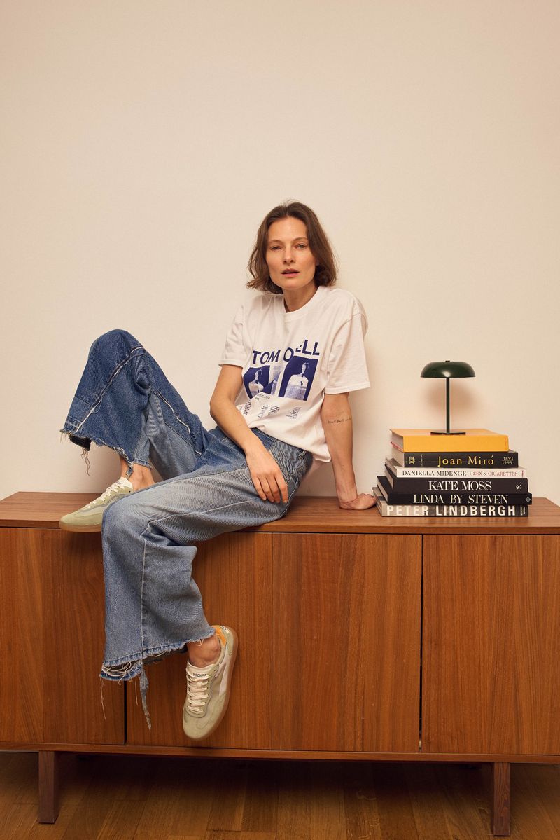 A young adult woman sits on a wooden cabinet in a bright room wearing jeans, sneakers and a graphic t-shirt with books and a lamp.