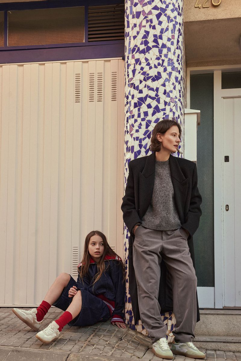 Model Tanya Ruban standing outdoors next to a mosaic column wearing a black coat, grey trousers, and sneakers.