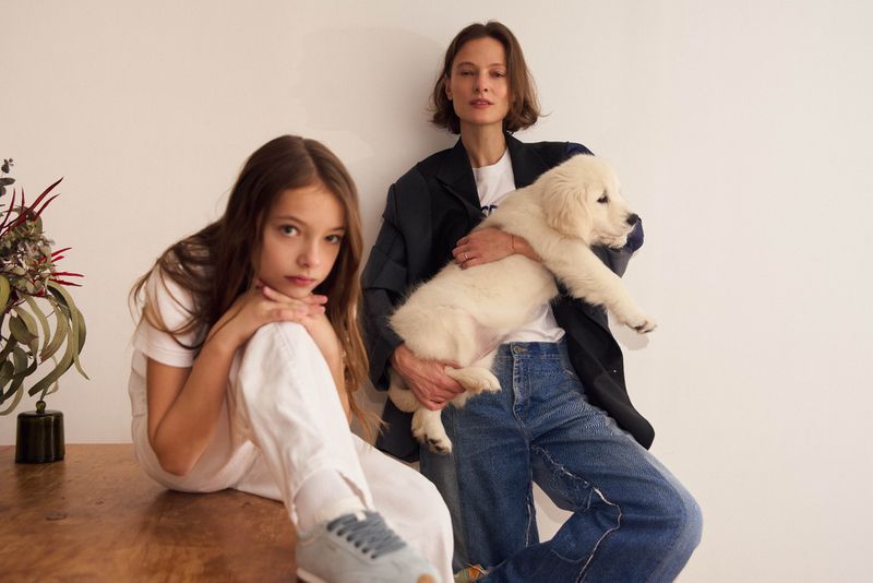 A mother in a blazer holds a Golden Retriever puppy while posing next to a young girl on a wooden table against a plain white backdrop.
