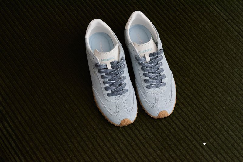 A pair of light blue suede sneakers with gray laces is displayed on a dark green corduroy surface, showcasing a minimalist aesthetic.