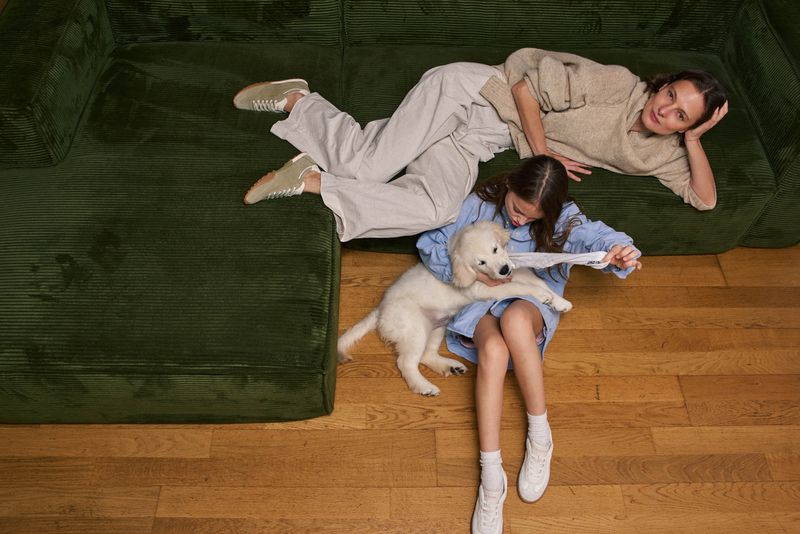 Indoor lifestyle shot of a mother lying on a green couch, watching her daughter play with a white puppy on a wooden floor.