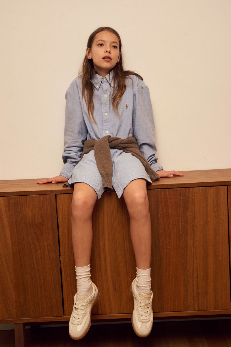 Girl in button-down shirt and sneakers sitting on a wooden cabinet in a brightly lit room, looking slightly off-camera.