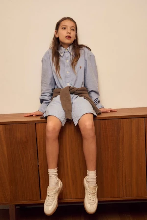 Girl in button-down shirt and sneakers sitting on a wooden cabinet in a brightly lit room, looking slightly off-camera.