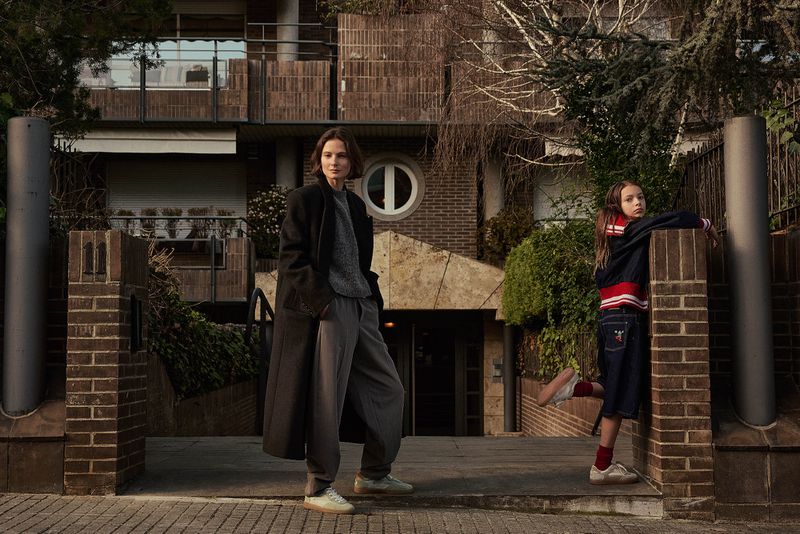 A woman and child standing outside a brick building. The woman is wearing a long coat and the child is wearing a bomber jacket.