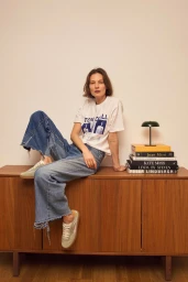 Model sitting on a wooden sideboard wearing a printed white t-shirt, wide-leg denim jeans, and sneakers for HOFF.