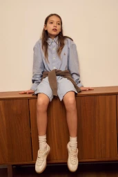 Young model Tanya Ruban seated on a wooden cabinet wearing a light blue button-down shirt, white socks, and sneakers for HOFF.