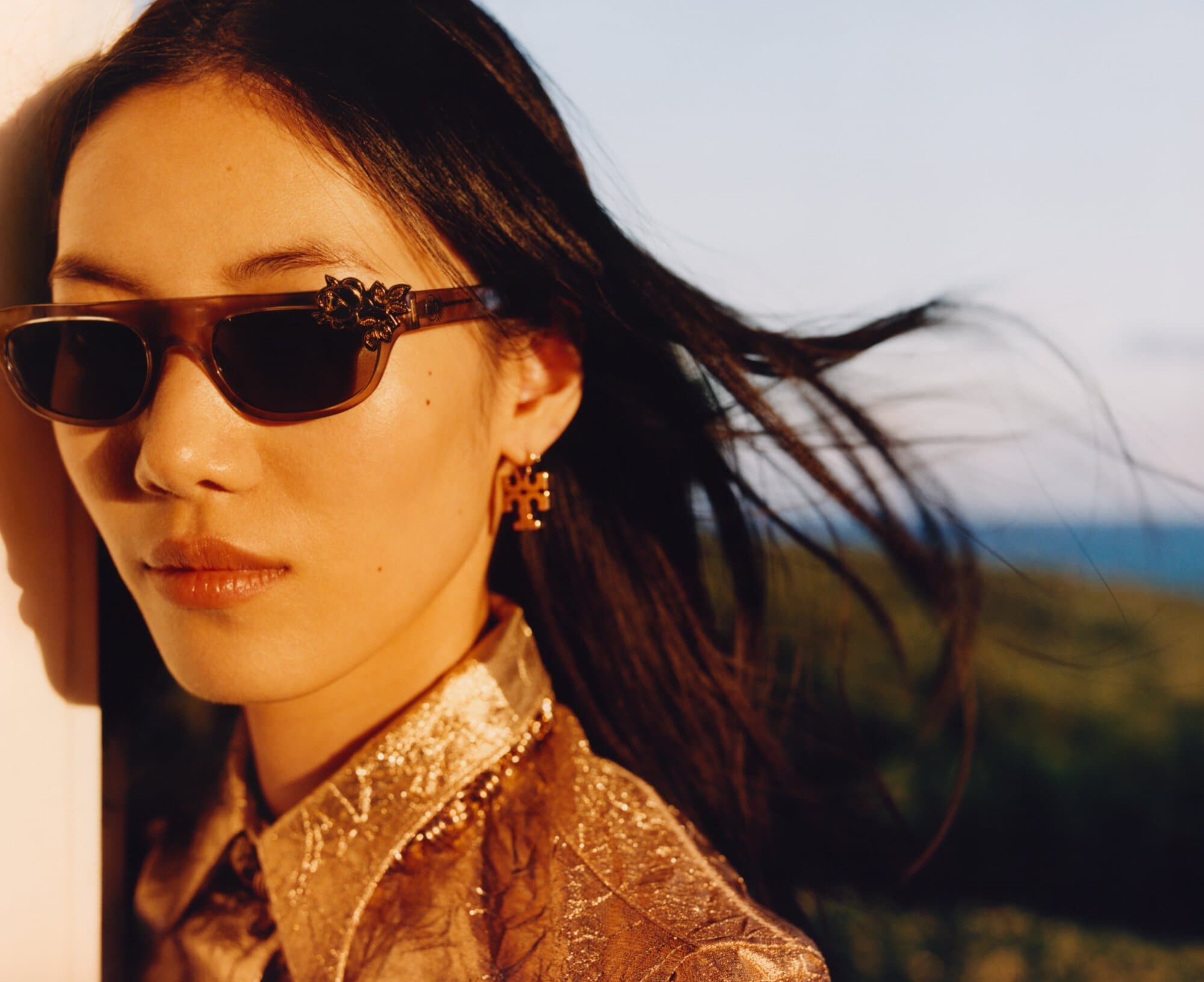 Portrait of an Asian woman wearing brown sunglasses with floral embellishments, gold earrings, and a shiny gold shirt outdoors.