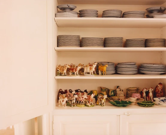 A shelving unit displays stacks of white plates and decorative figurines of cows in an indoor environment with a neutral color palette.