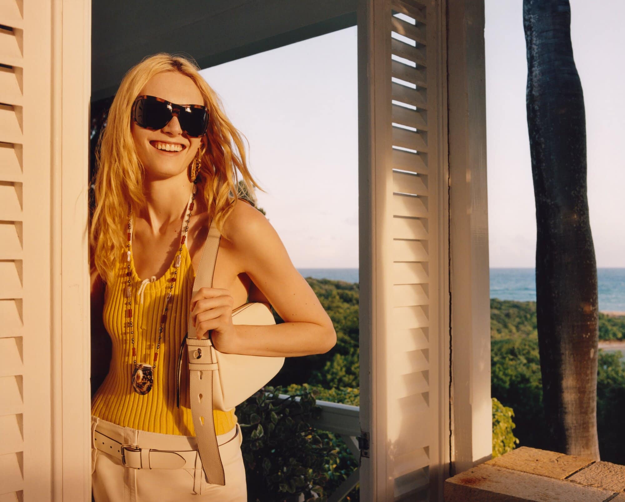 Blonde woman smiles on a veranda overlooking the ocean wearing sunglasses, a yellow top, white skirt, and a white handbag.