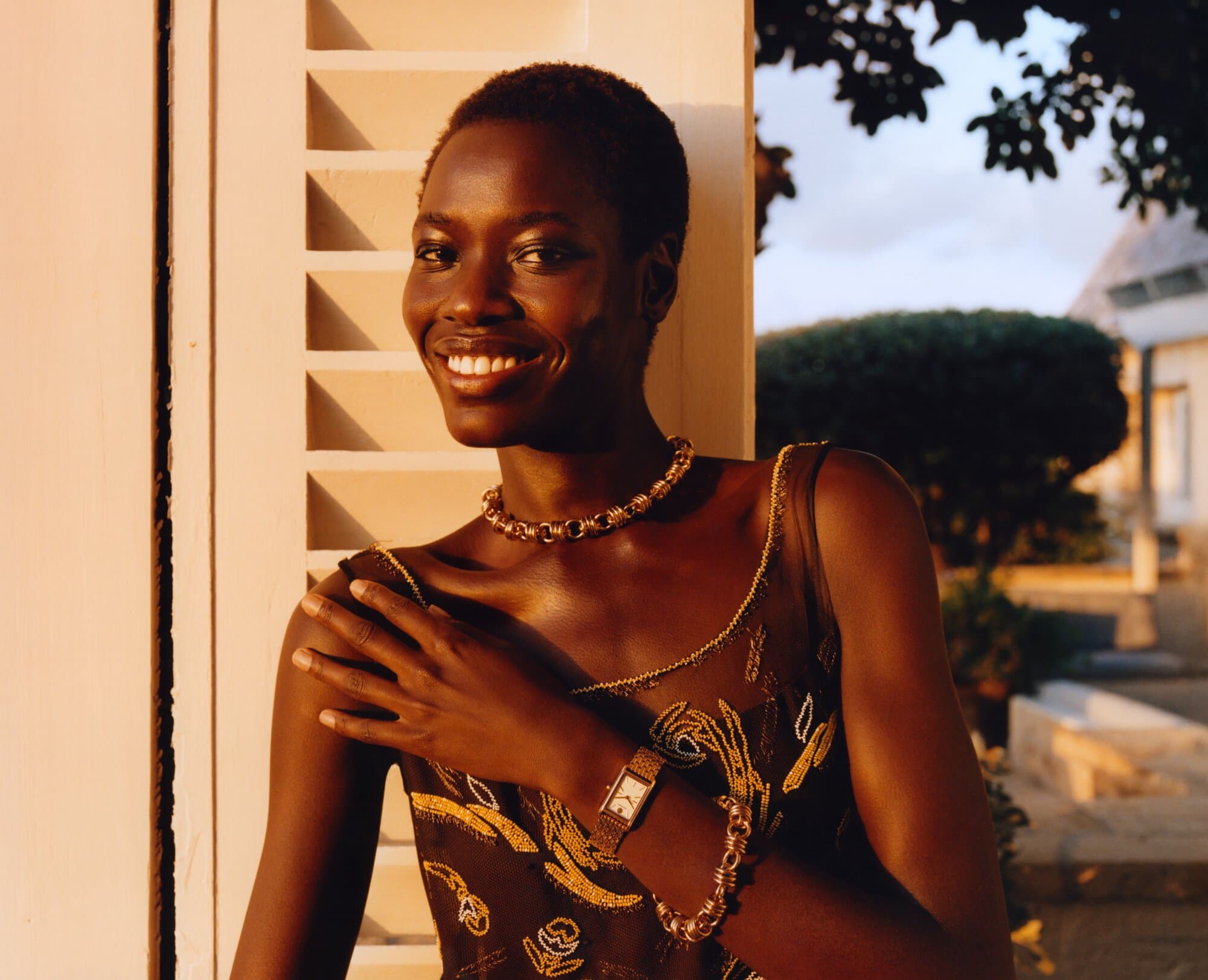 Smiling Black woman poses next to a shuttered white window wearing a dark floral dress, gold jewelry, and wristwatch outside during daytime.