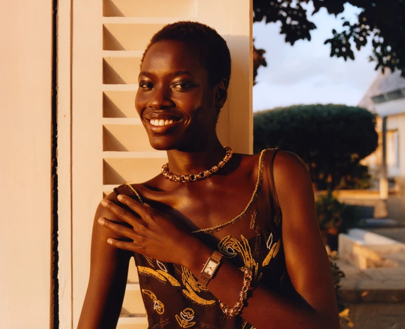 Smiling Black woman poses next to a shuttered white window wearing a dark floral dress, gold jewelry, and wristwatch outside during daytime.