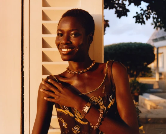 Smiling Black woman poses next to a shuttered white window wearing a dark floral dress, gold jewelry, and wristwatch outside during daytime.