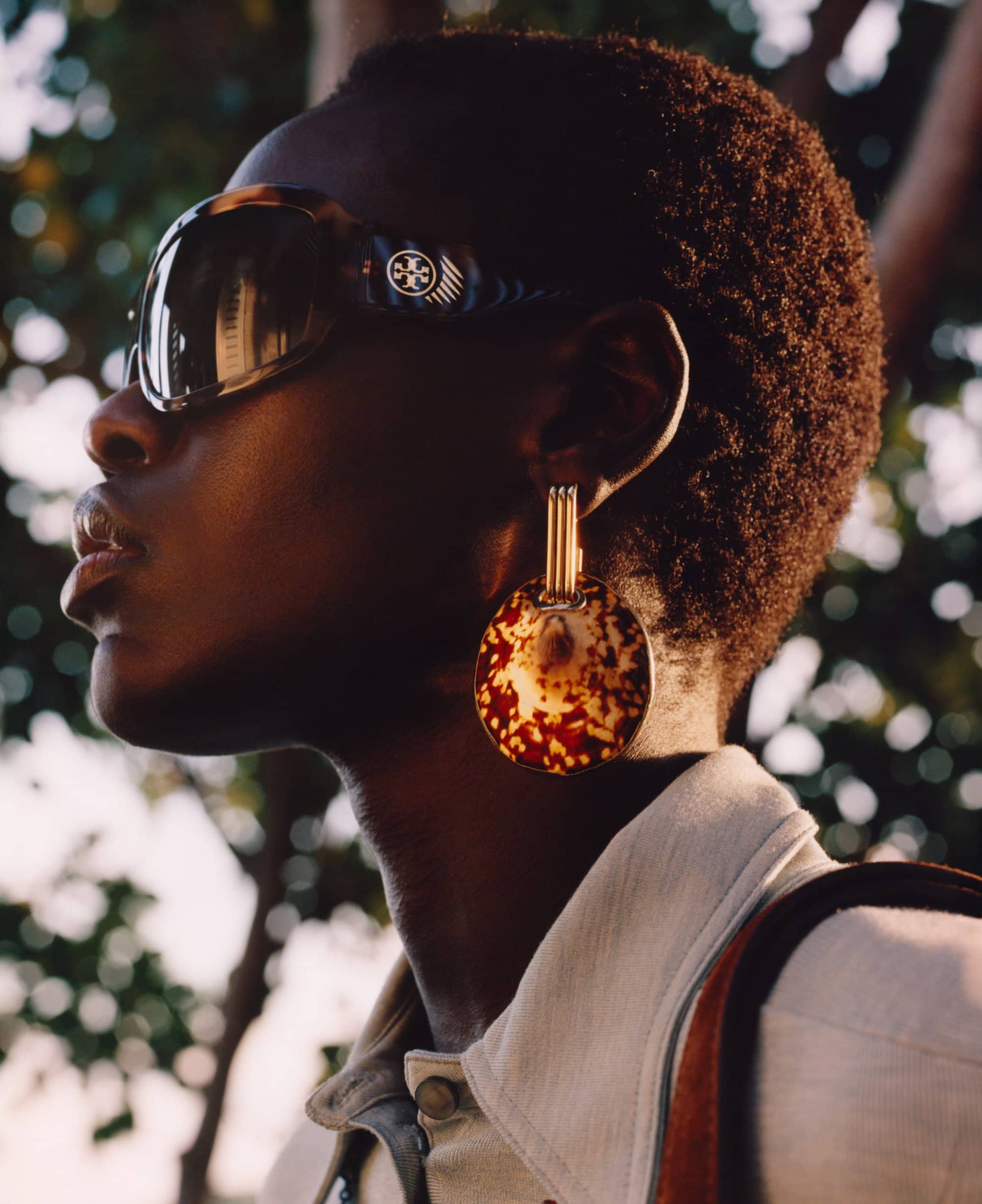 A dark-skinned woman with an Afro, wearing sunglasses and large earrings, poses outdoors with blurred green foliage in the background.