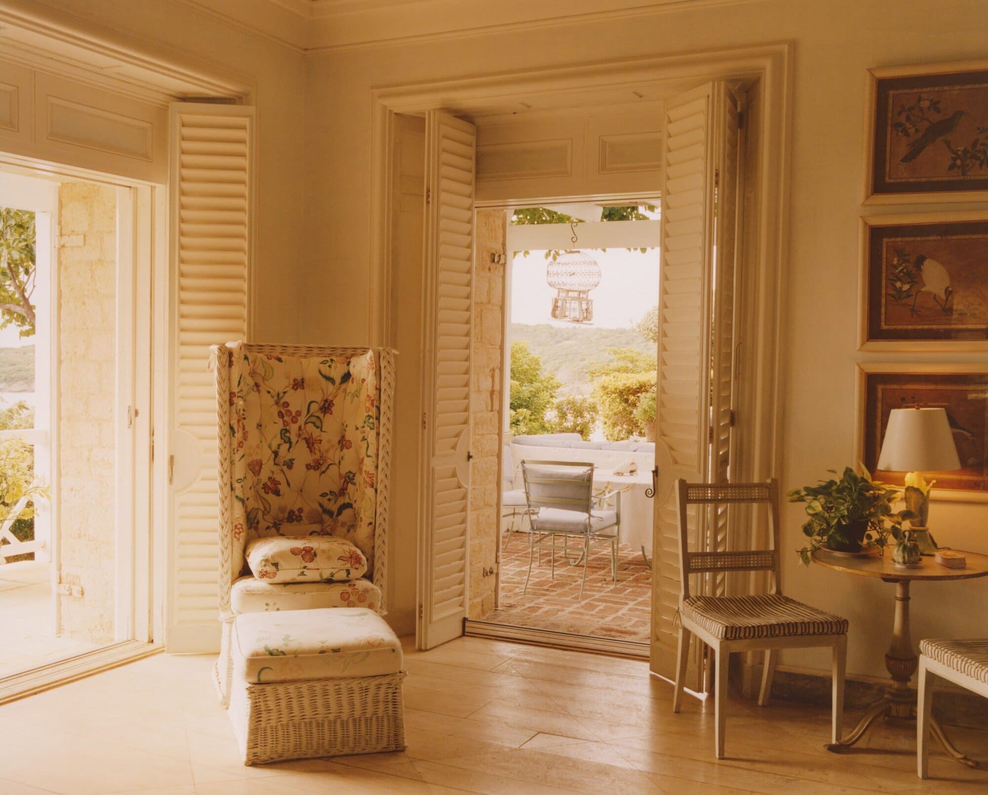 Bright interior with floral-patterned chair, shutters, artwork, and open patio view in soft lighting showcases classic and elegant design.