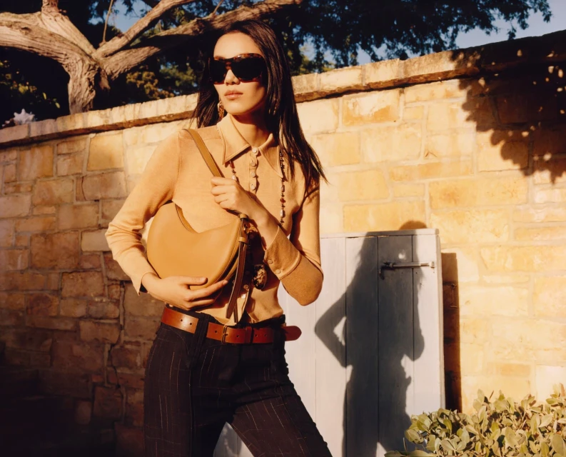 Stylish woman in tan top, striped pants, sunglasses posing outdoors with a leather shoulder bag against a brick wall backdrop.