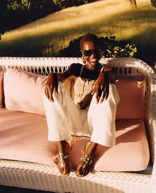 Smiling Black woman with short hair sitting cross-legged on a wicker couch with pink cushions in an outdoor setting with greenery.