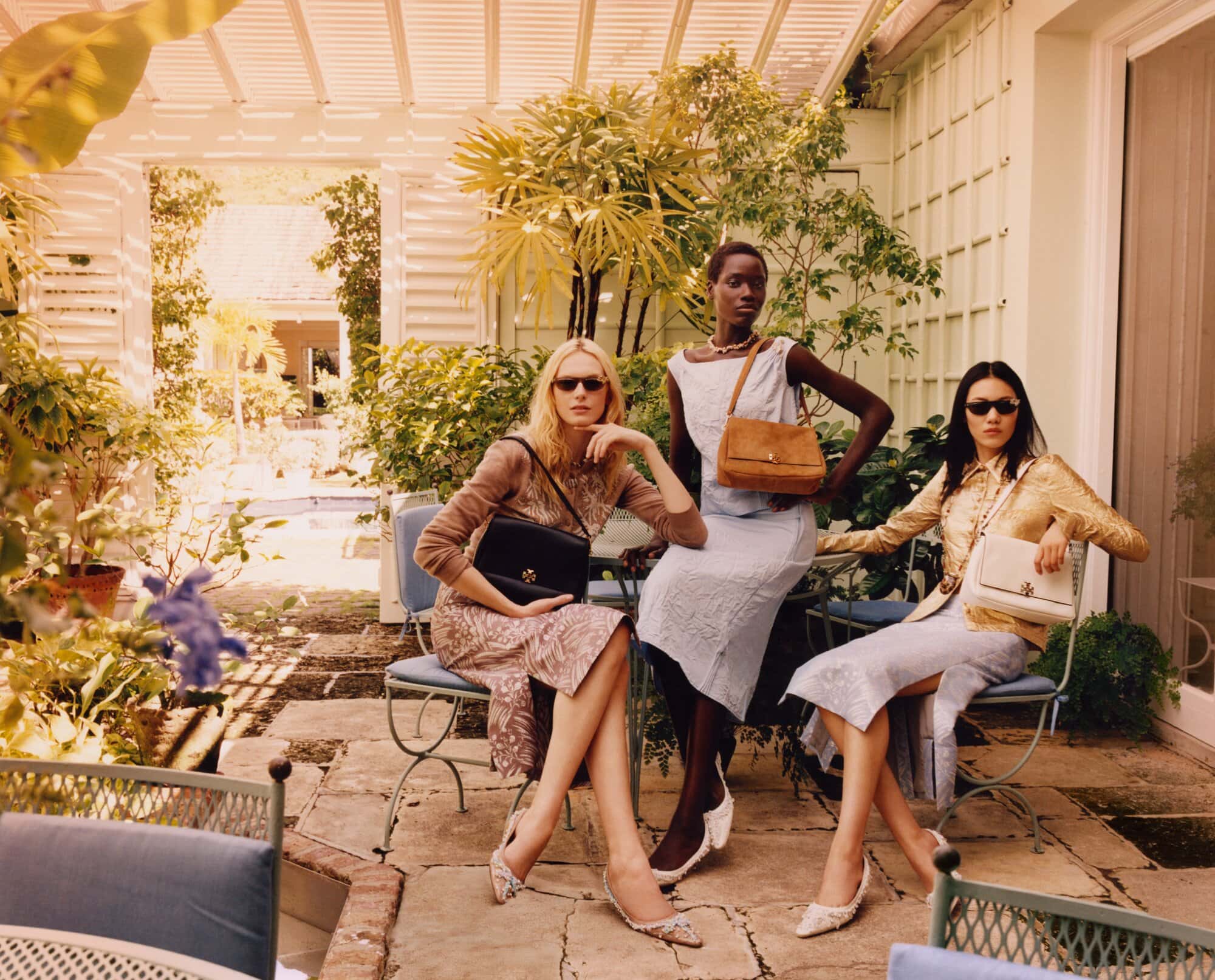 Three diverse women showcase purses outdoors. The model in the center stands while two others sit on blue metal chairs in sunlight.