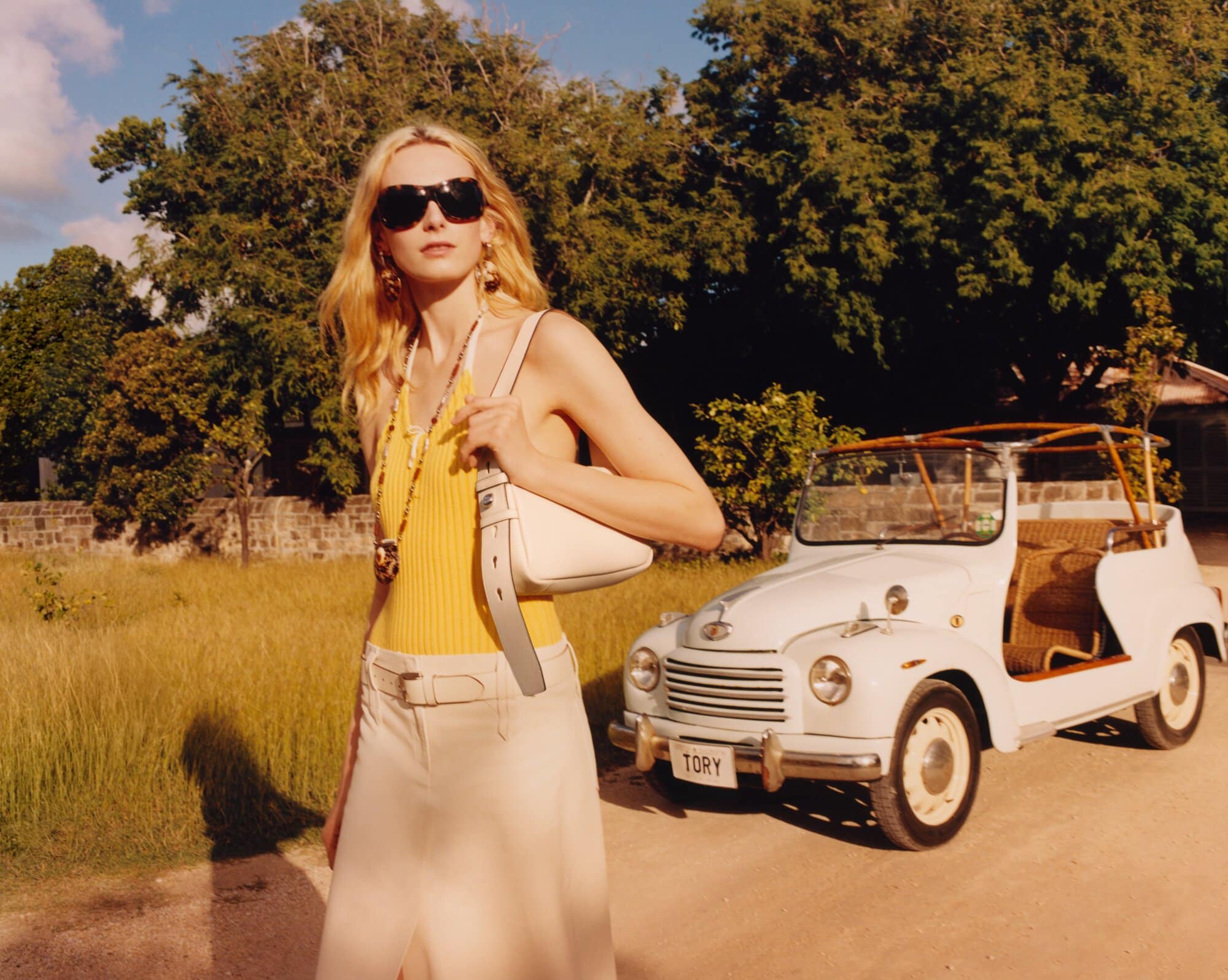 Fashion shoot featuring a blonde model in a yellow top and skirt, sunglasses, and handbag, posing beside a vintage car outdoors.