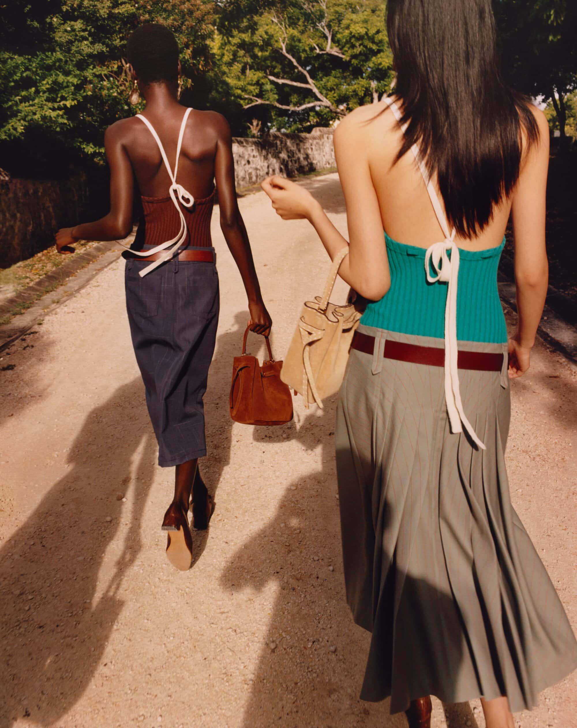 Two women stroll down a tree-lined path, carrying stylish handbags in muted earth tones, captured in an outdoor fashion shot.