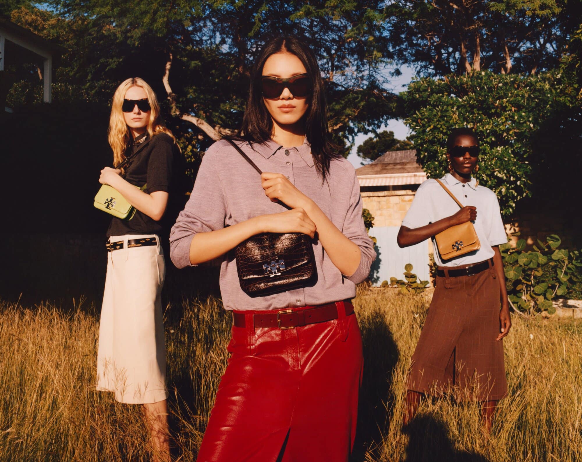 Three fashion models pose outdoors in the sunlight, showcasing purses. The background features a home, trees, and tall grass.