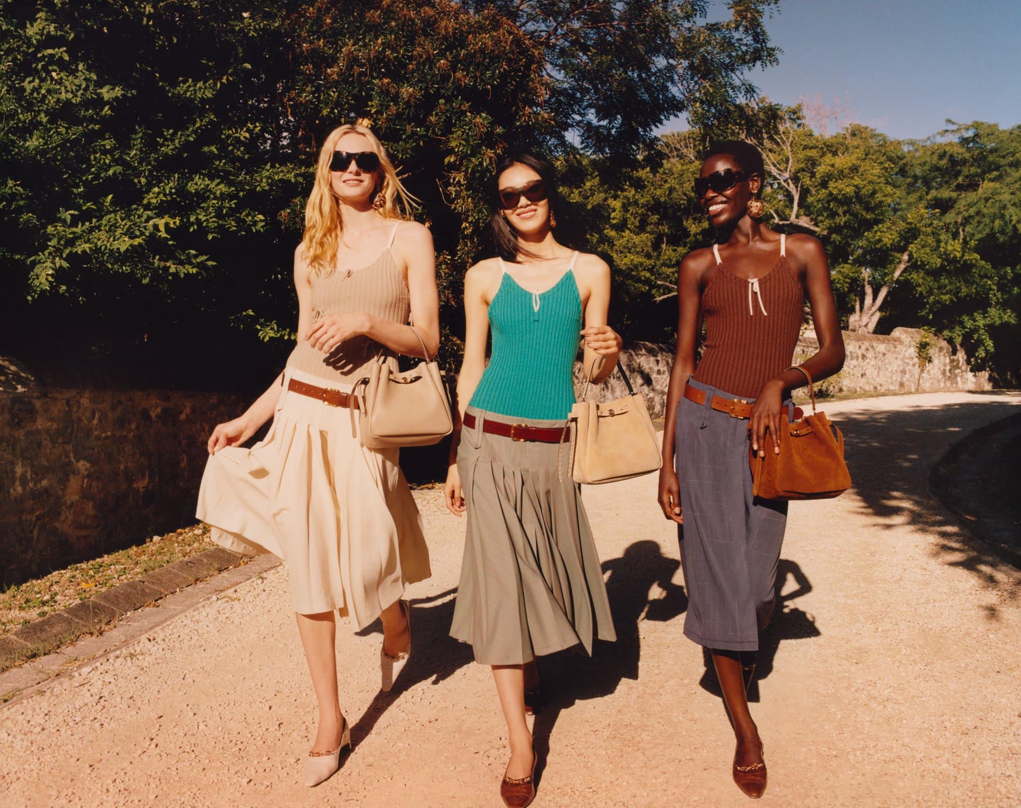 Three fashion models wearing skirts, tank tops, and sunglasses walk along a sunlit path with trees in the background, each carrying a handbag.