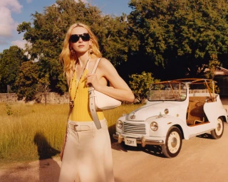 Model wearing dark sunglasses and a yellow top standing outdoors next to a small white vintage car