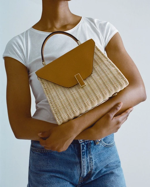 Woman in denim shorts holds a wicker and brown leather handbag in a studio setting with neutral lighting and soft, diffused light.