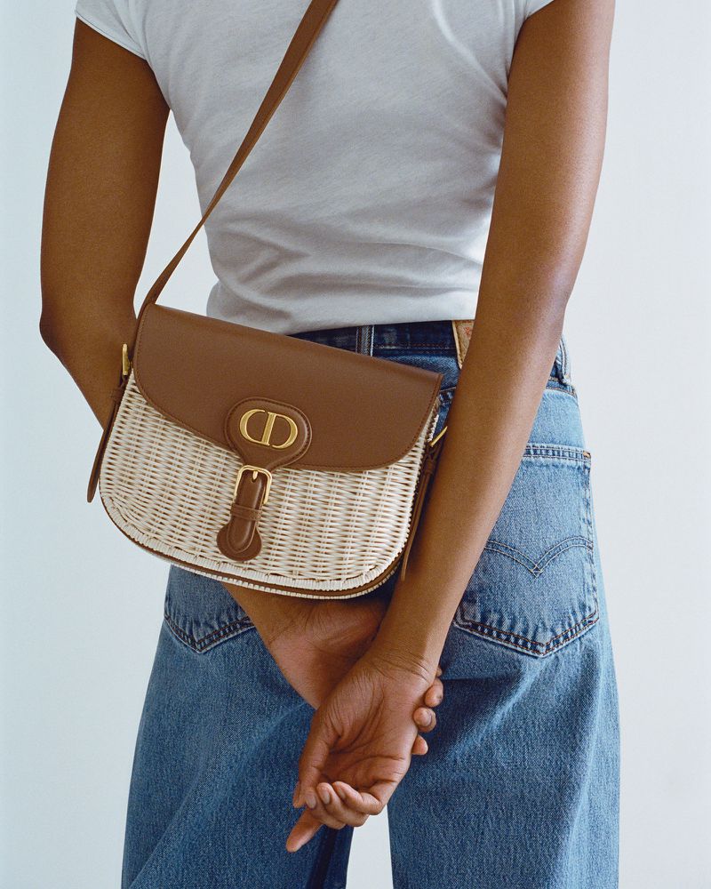 Woman wearing a white t-shirt and blue jeans with a brown and white woven crossbody bag with a gold logo buckle and hardware.