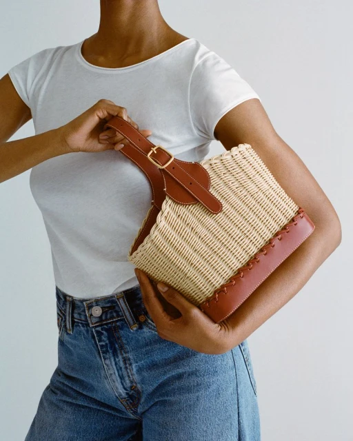 A person holds a straw tote bag with brown leather accents, wearing a white t-shirt and jeans against a plain background.