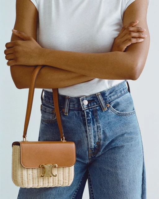 Tan-and-wicker Celine bag hanging from the shoulder of an adult woman wearing a white t-shirt and blue jeans against a white backdrop.