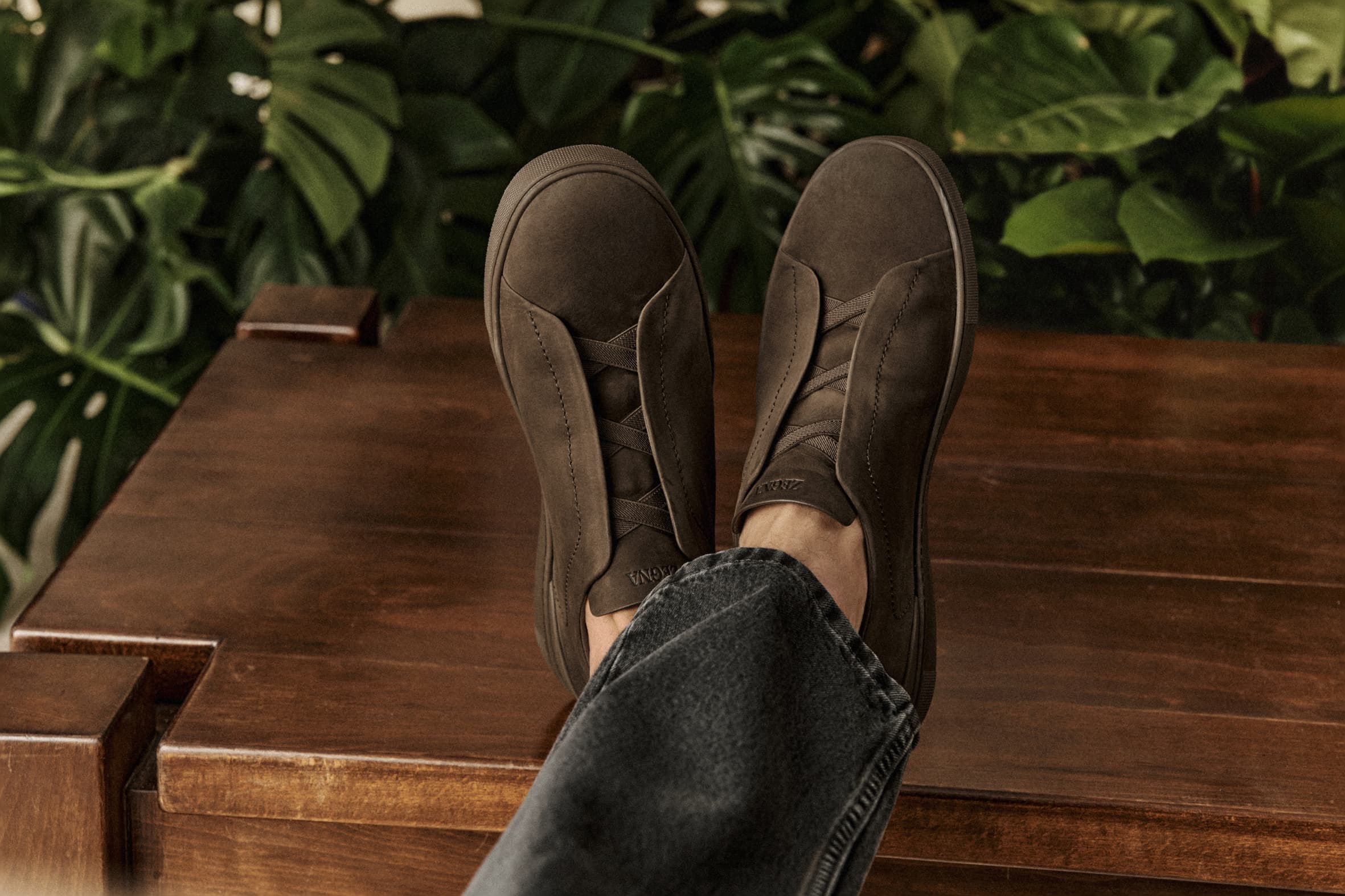 Close-up of brown suede slip-on sneakers with dark-wash denim jeans and a wood surface; indoor setting with green foliage backdrop.