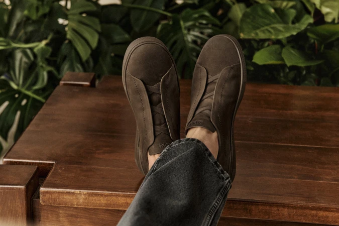 Close-up of brown suede slip-on sneakers with dark-wash denim jeans and a wood surface; indoor setting with green foliage backdrop.