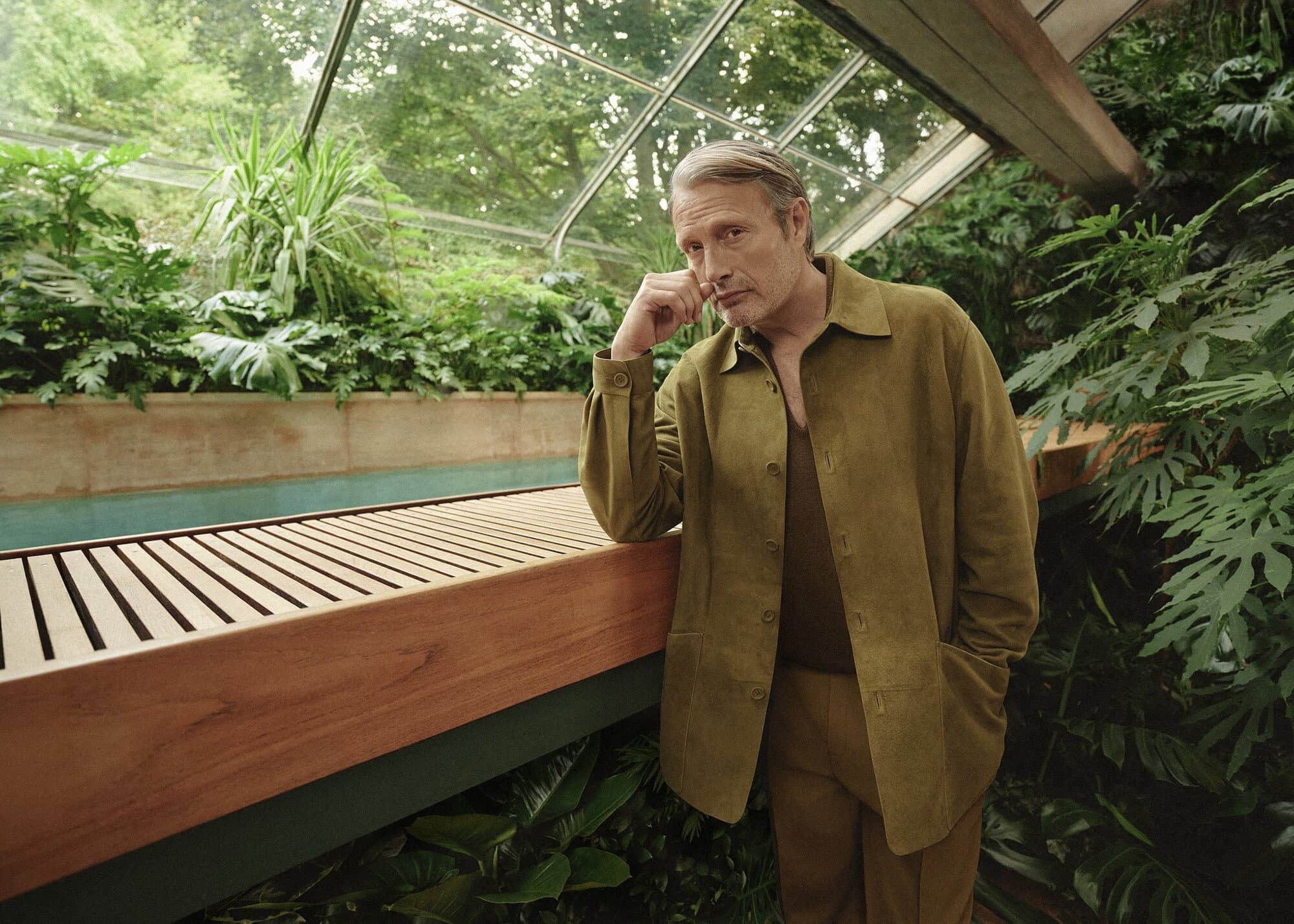 Adult male in tan shirt and jacket leans against a wooden structure in a greenhouse surrounded by lush green plants and natural light.