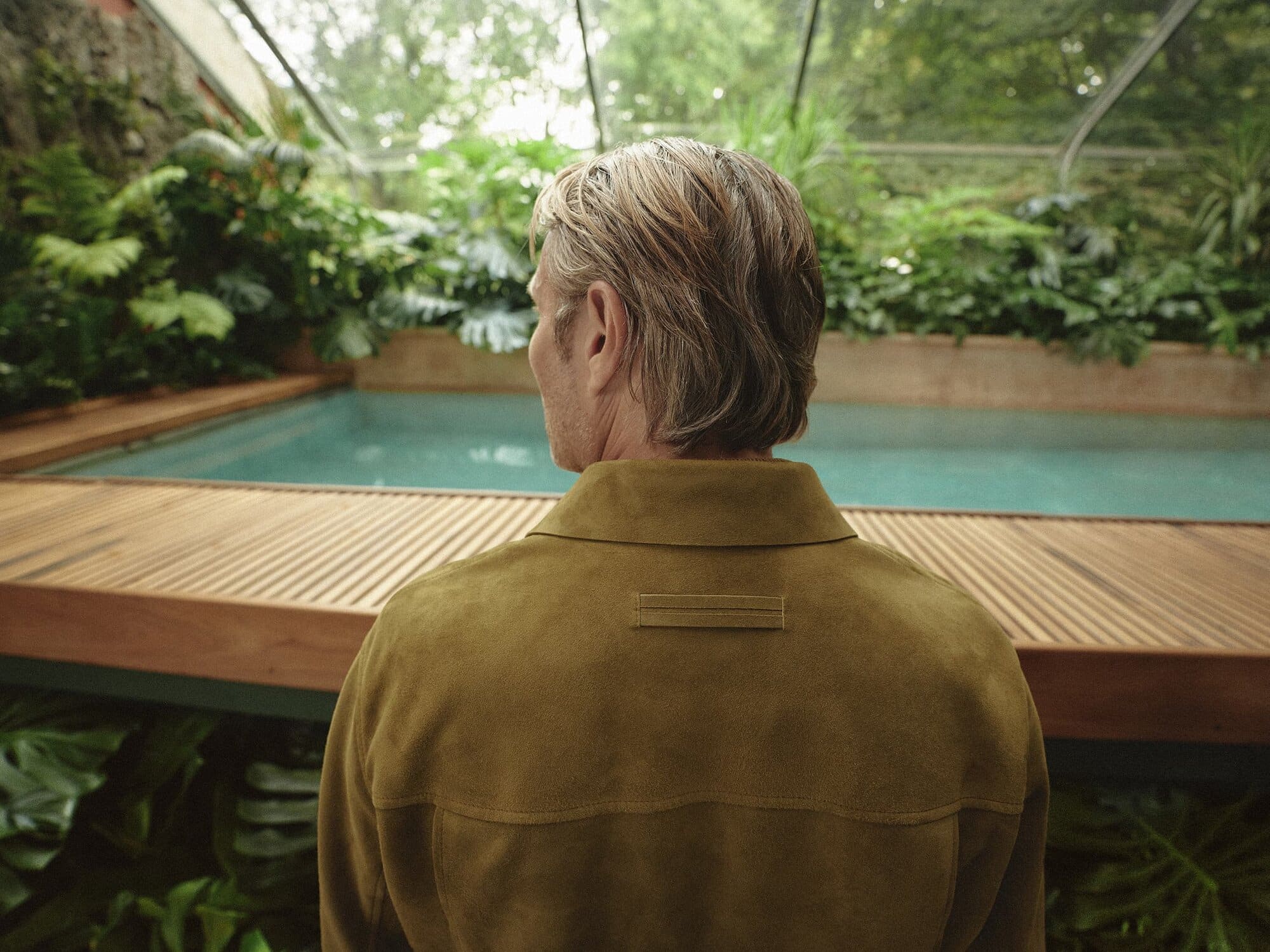 Man in a tan jacket contemplates a turquoise swimming pool surrounded by lush tropical foliage inside of a bright glass enclosure.