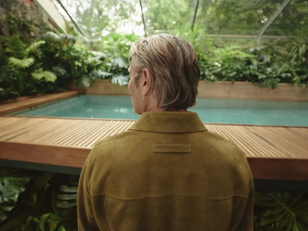 Man in a tan jacket contemplates a turquoise swimming pool surrounded by lush tropical foliage inside of a bright glass enclosure.