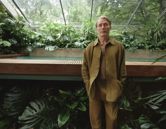 Adult male in a tan suit jacket standing indoors amid lush green plants and ferns, inside a glass-enclosed sunroom or greenhouse.