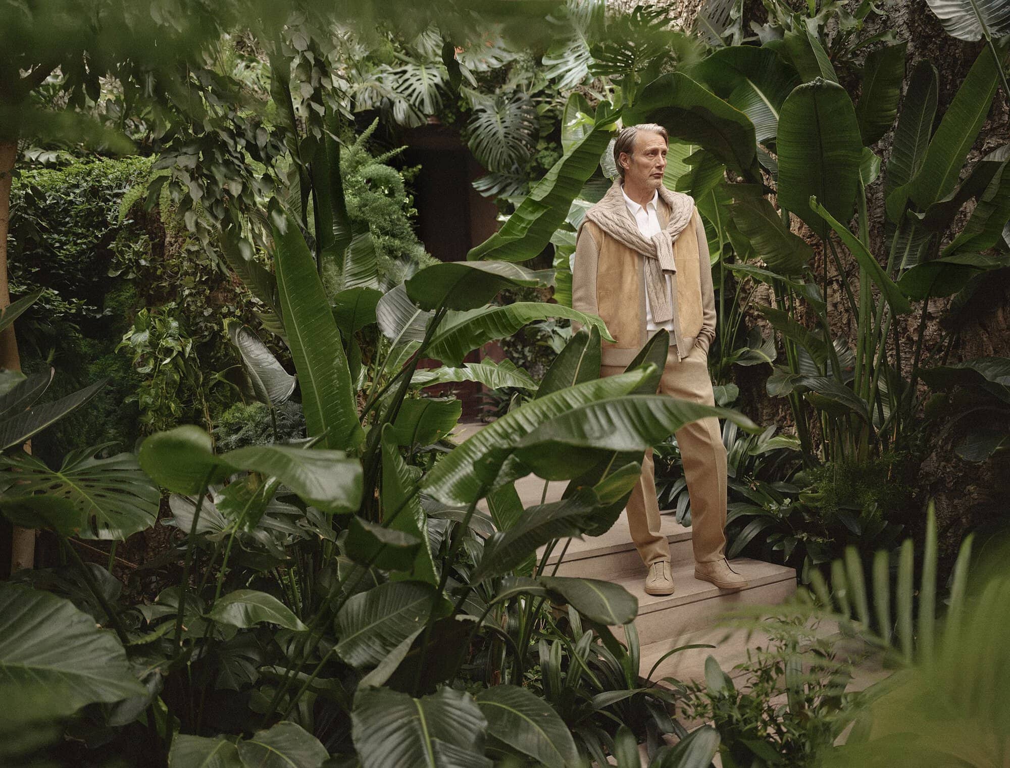 Adult man in tan suit and scarf stands among lush green tropical plants and stairs outdoors in natural light, looking to the side.