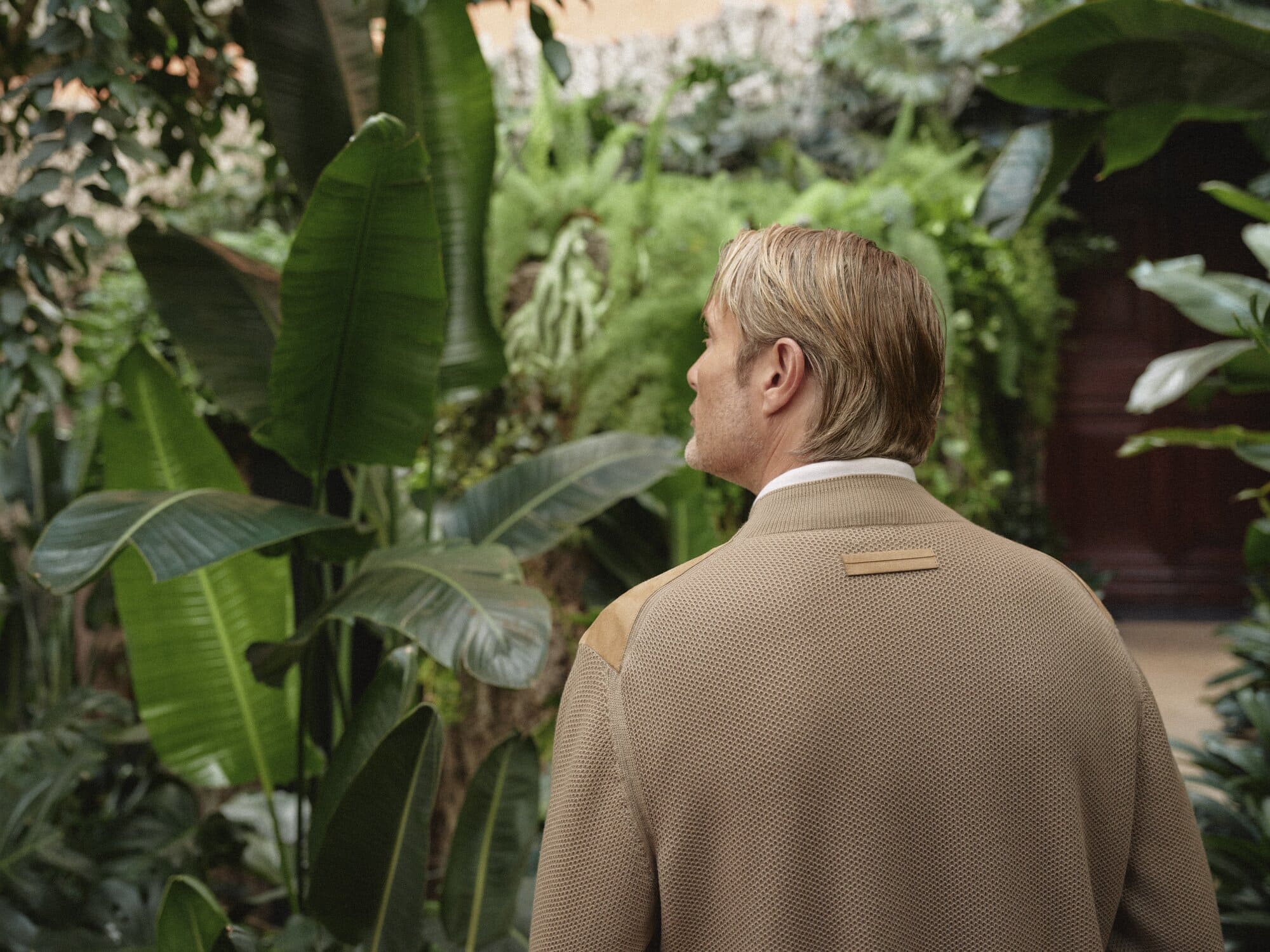 Adult male with blond hair stands in front of a lush green wall, wearing a tan collared shirt with tan suede shoulder pads.